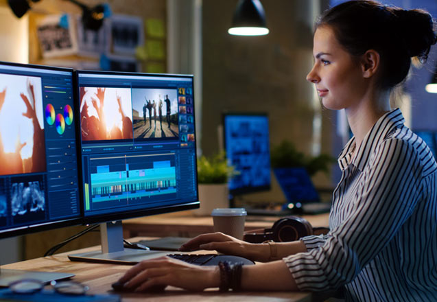 A young female video editor works on her latest project while looking at 2 side-by-side monitors